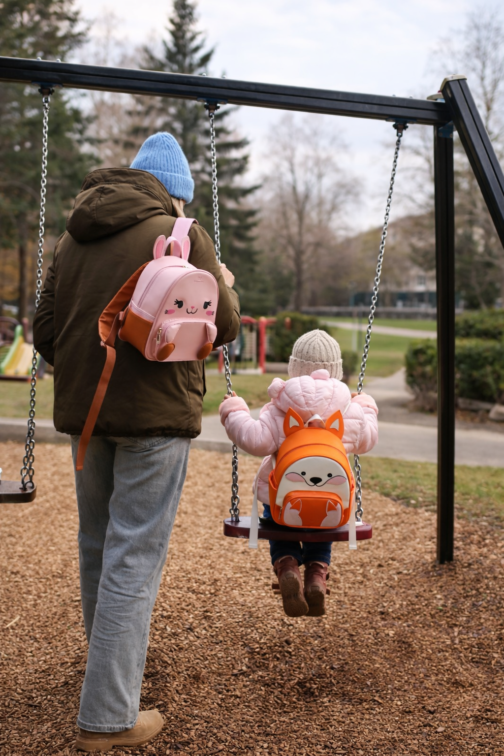 Two children on swings at a playground with backpacks.